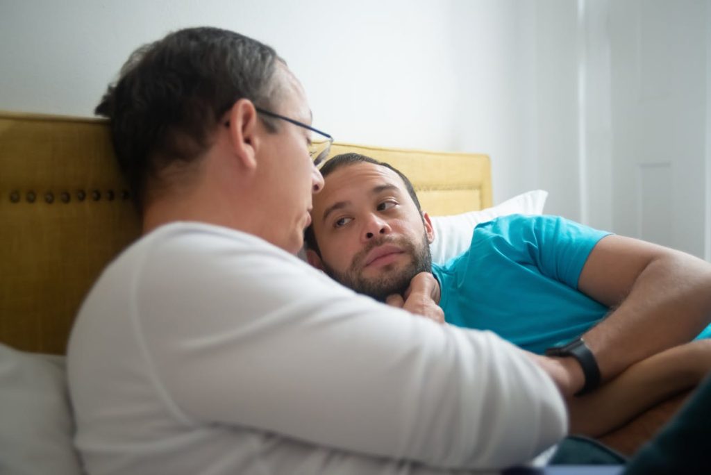 Gay couple lying in bed deep conversation about open relationships.
