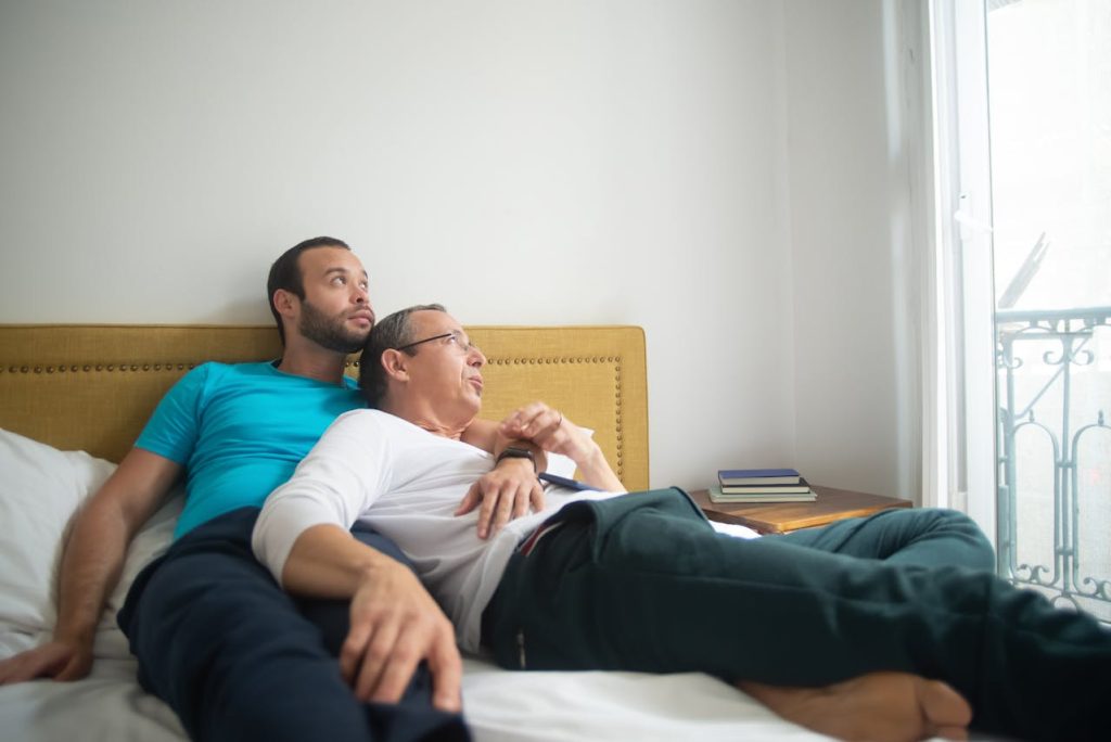 Two adult men, one older and one younger, lie embracing in bed. They are fully clothed and both looking out a window in the same direction, symbolising the shared relationship that might be termed daddy issues for the younger partner