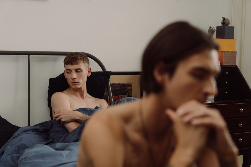 A couple on a bed after a fight, both appearing unhappy, representing that gay domestic violence can show up as repeated demands and abuse.