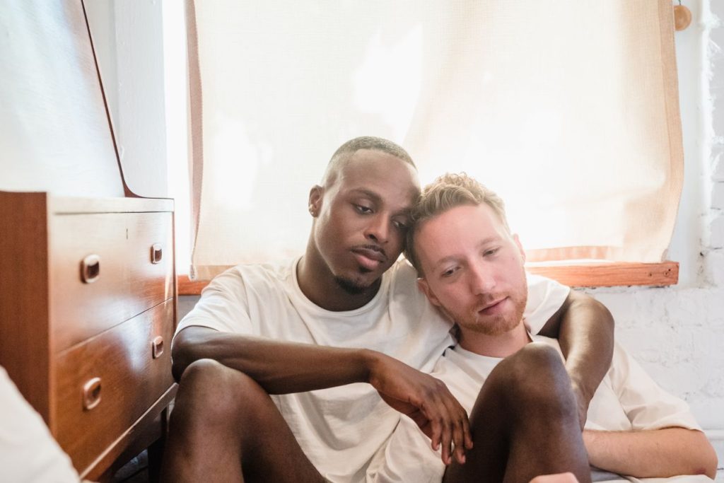 A sad gay couple sitting on the floor under a window and beside a chest of drawers in the aftermath of gay domestic violence